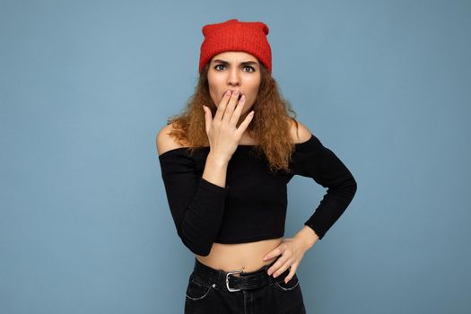 Young emotional surprised shocked nice winsome brunet woman wavy-haired with sincere emotions wearing black crop top and red hat isolated on blue background with free space.