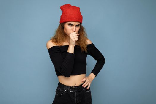 Shot of young emotional angry nice winsome brunet woman wavy-haired with sincere emotions wearing black crop top and red hat isolated on blue background with free space and holding hand near mouth.