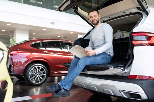a buyer studying a booklet in a showroom of a car dealership sitting in the trunk of a crossover looking at the camera with a smile.