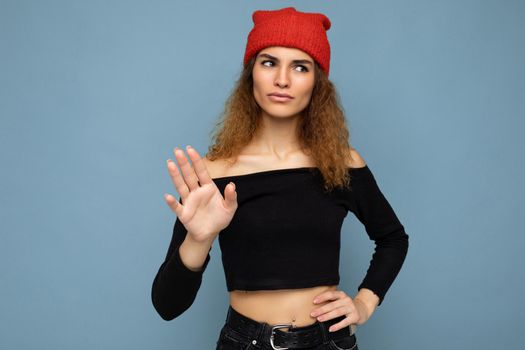 Shot of young self-confident nice winsome brunet woman wavy-haired with sincere emotions wearing black crop top and red hat isolated on blue background with free space and showing stop gesture.