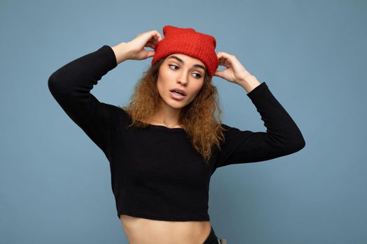 Young pretty brunette woman curly with sincere emotions wearing stylish black crop top and red hat isolated on blue background with copy space.