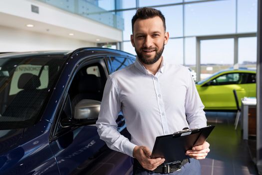 charming salesman in a car dealership against the background of new premium cars.