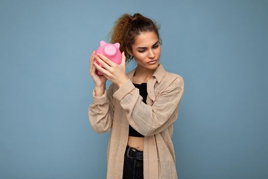 Photo of sincere sad cute young beautiful attractive woman with dark curly hair in a ponytail wearing casual beige shirt isolated over blue background with copy space and holds pink piggy box. Moneybox concept.