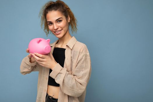 Portrait photo of happy positive smiling young winsome attractive brunette curly woman with sincere emotions wearing stylish beige shirt isolated over blue background with copy space and holding pink piggy box. Money box concept.