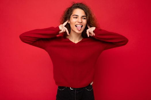 Photo of young positive happy joyful attractive brunette wavy-haired woman with sincere emotions wearing casual red sweater isolated on red background with empty space and showing tongue.