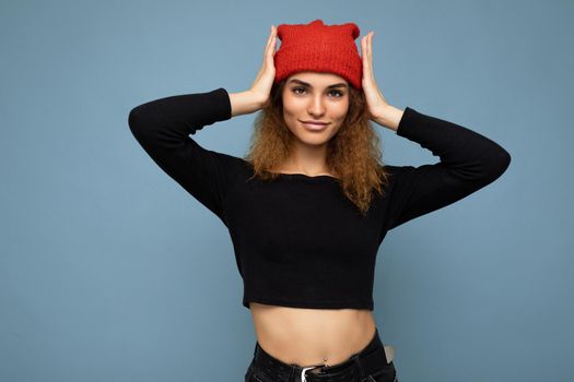 Photo of young pretty positive brunette woman curly with sincere emotions wearing stylish black crop top and red hat isolated on blue background with copy space.