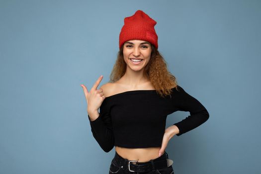 Photo of young positive happy beautiful brunette wavy-haired woman with sincere emotions wearing black crop top and red hat isolated on blue background with copy space.