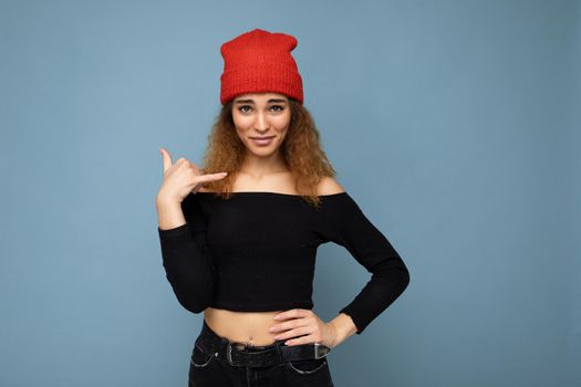 Photo of young positive beautiful brunette curly woman with sincere emotions wearing black crop top and red hat isolated on blue background with copy space and showing phone gesture.