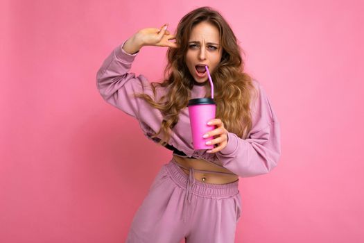 Photo shot of young beautiful attractive cute positive happy smiling curly blonde woman wearing trendy and stylish pink sport suit isolated over pink background with copy space for text and holding cup of milkshake. Summertime concept.