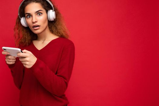 Photo of beautiful young brunette curly shocked amazed woman wearing dark red sweater isolated over red background wall with free space wearing white bluetooth headphones listening to music and holding mobile phone.