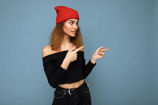 Photo of young attractive brunette curly woman with sincere emotions wearing black crop top and red hat isolated on blue background with empty space and pointing at copy space.