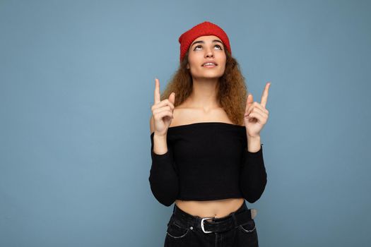 Photo of young attractive brunette curly woman with sincere emotions wearing black crop top and red hat isolated on blue background with empty space and pointing up at copy space.