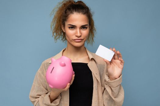 Young dissatisfied sad upset winsome attractive brunette curly woman with sincere emotions wearing stylish clothes isolated on blue background with copy space, holds pink piggy box and credit card for shopping.