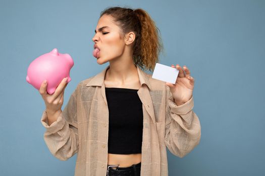 Photo of cute angry funny young beautiful attractive woman with dark curly hair in a ponytail wearing casual beige shirt isolated over blue background with copy space and holds pink piggy box for coins and white bank card for mockup.