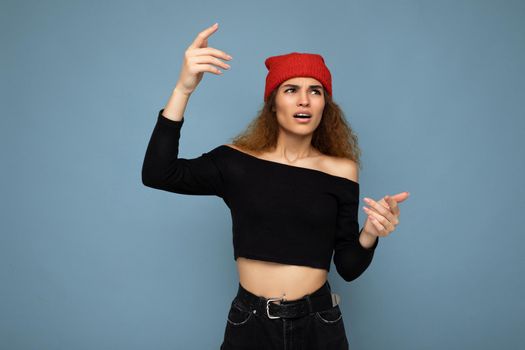 Young emotional dissatisfied pretty brunette woman wavy-haired with sincere emotions wearing black crop top and red hat isolated on blue background with free space.