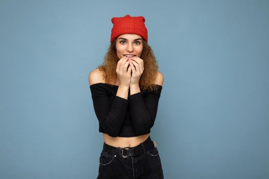 Shot of young emotional nice winsome brunet woman wavy-haired with sincere emotions wearing black crop top and red hat isolated on blue background with free space.