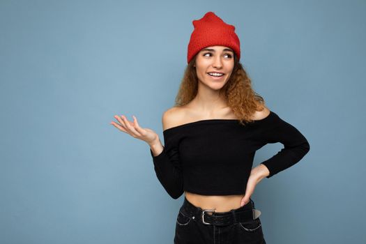 Photo of young positive happy beautiful brunette wavy-haired woman with sincere emotions wearing black crop top and red hat isolated on blue background with copy space.
