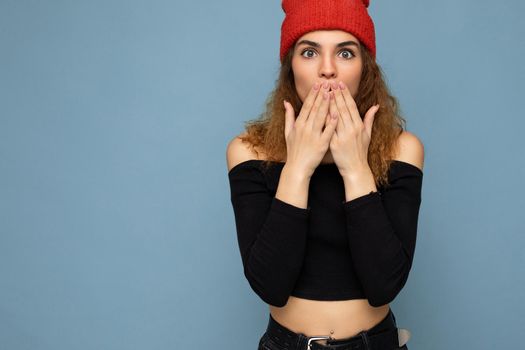 Shot of young emotional astonished nice winsome brunet woman wavy-haired with sincere emotions wearing black crop top and red hat isolated on blue background with free space and covering mouth.