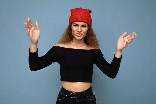 Young emotional dissatisfied pretty brunette woman wavy-haired with sincere emotions wearing black crop top and red hat isolated on blue background with free space and having questions.
