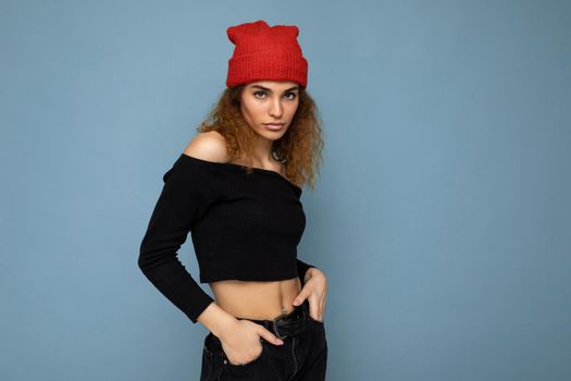 Photo of young positive self-confident cute nice brunette woman curly with sincere emotions wearing stylish black crop top and red hat isolated on blue background with copy space.