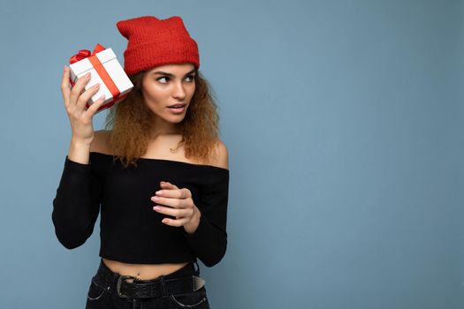 Photo shot of beautiful thoughtful young dark blonde curly woman isolated over blue background wall wearing black crop top and red hat holding gift box looking to the side trying to guess the present in the box. Copy space