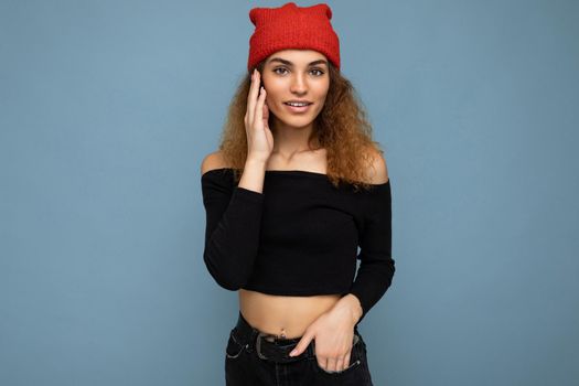 Photo of pretty beautiful attractive cute positive happy cool young curly dark blonde woman standing isolated over blue background wall with copy space for text wearing everyday trendy and stylish black top and red hat.