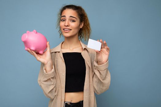 Portrait photo of happy positive smiling young charming attractive brunette curly woman with sincere emotions wearing stylish beige shirt isolated over blue background with empty space, holds pink piggy box and credit card. Shopping concept.