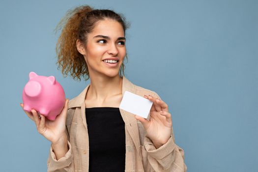 Photo of happy positive smiling young fascinating nice brunette curly woman with sincere emotions wearing stylish beige shirt isolated over blue wall with copy space, holds pink piggy box and credit card. Shopping concept.
