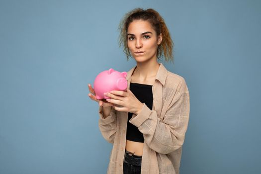 Portrait photo of sincere cute young charming beautiful attractive woman with dark curly hair in a ponytail wearing casual beige shirt isolated over blue background with copy space and holds pink piggy box. Moneybox concept.