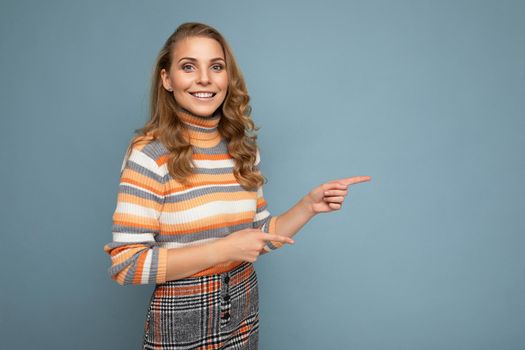 Portrait photo of young winsome beautiful attractive happy smiling blonde woman with wavy-hair wearing stylish striped sweater isolated over blue background and showing with fingers to empty space for text.