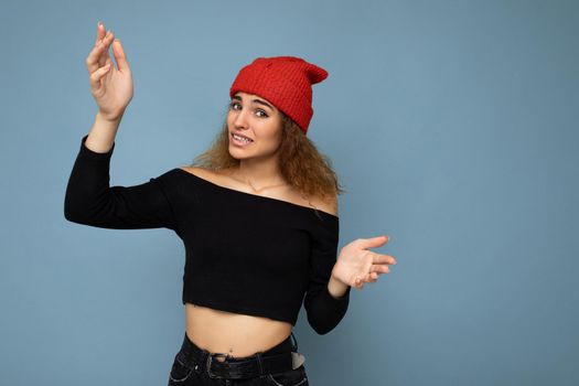 Young emotional dissatisfied funny pretty brunette woman wavy-haired with sincere emotions wearing black crop top and red hat isolated on blue background with free space and explaining something.