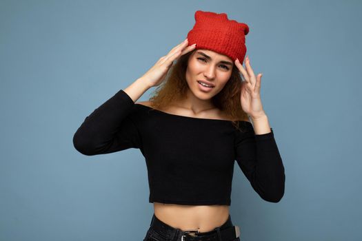 Young emotional dissatisfied pretty brunette woman curly with sincere emotions wearing stylish black crop top and red hat isolated on blue background with copy space and have headache. Pain concept.