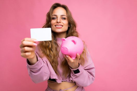 Photo shot of young beautiful attractive cute positive happy smiling curly blonde woman wearing trendy and stylish pink sport suit isolated over pink background with copy space for text and holding pink penny pig and white bank plastic card for money.