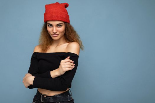 Photo of beautiful self-confident sexy young brunette curly woman isolated over blue background wall wearing black crop top and red hat looking at camera. Copy space