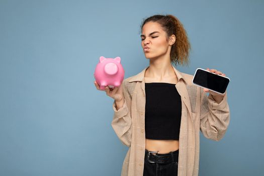 Portrait photo of dissatisfied doubtful young beautiful attractive brunette curly woman with sincere emotions wearing stylish beige shirt isolated over blue background with copy space, holds pink piggy box and shows mobile phone with empty screen display. Payment concept.