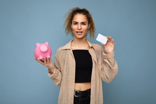Young dissatisfied asking winsome attractive brunette curly woman with sincere emotions wearing stylish clothes isolated on blue background with copy space, holds pink piggy box and credit card for shopping.