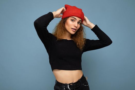 Young pretty positive brunette woman curly with sincere emotions wearing stylish black crop top and red hat isolated on blue background with copy space.