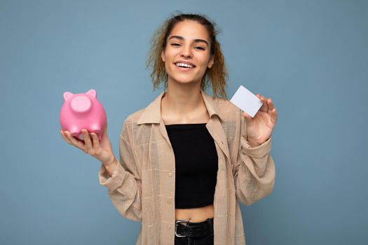 Portrait photo of happy positive smiling young beautiful attractive brunette curly woman with sincere emotions wearing stylish beige shirt isolated over blue background with copy space, holds pink piggy box and credit card. Shopping concept.