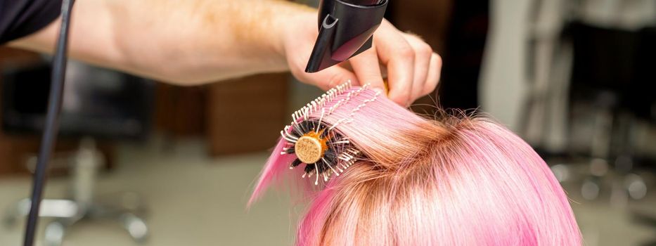 Drying short pink hair of young caucasian woman with a black hairdryer and black round brush by hands of a male hairdresser in a hair salon, close up