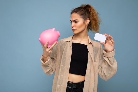 Portrait of young dissatisfied sad upset nice beautiful brunette curly woman with sincere emotions wearing stylish clothes isolated on blue background with copy space, holds pink pig moneybox and credit card for shopping.