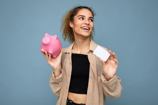 Portrait photo of happy positive smiling young beautiful attractive brunette curly woman with sincere emotions wearing stylish beige shirt isolated over blue background with copy space, holds pink pig penny bank and credit card. Shopping concept.