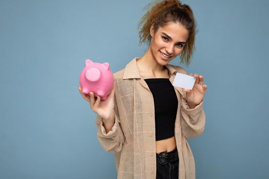 Portrait of happy positive smiling young beautiful attractive brunette curly woman with sincere emotions wearing stylish beige shirt isolated over blue wall with free space, holds pink pig coin bank and credit card. Shopping concept.
