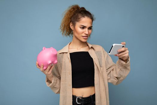Portrait photo of dissatisfied young beautiful attractive brunette curly woman with sincere emotions wearing stylish beige shirt isolated over blue background with copy space, holds pink piggy box and uses mobile phone. I don't understand.