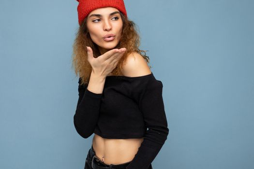 Photo of young positive cute nice brunette woman curly with sincere emotions wearing stylish black crop top and red hat isolated on blue background with copy space and giving kiss.