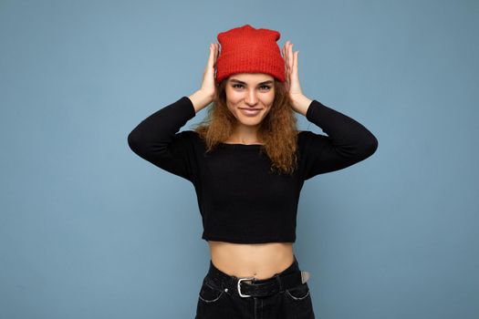 Shot of young nice winsome brunet woman wavy-haired with sincere emotions wearing black crop top and red hat isolated on blue background with free space and covering ears with hands.