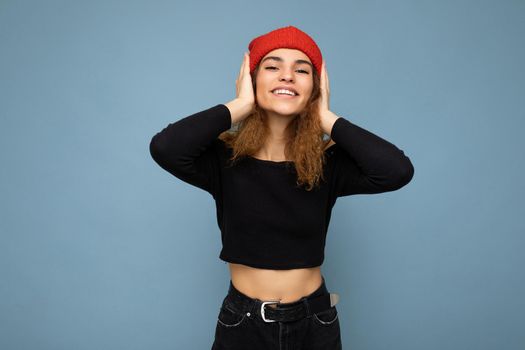 Shot of young positive smiling nice winsome brunet woman wavy-haired with sincere emotions wearing black crop top and red hat isolated on blue background with free space and covering ears.