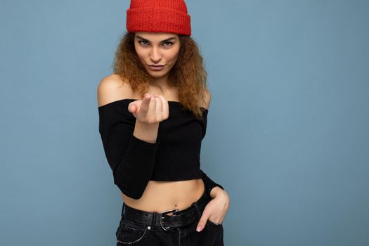 Photo of young positive cute nice brunette woman curly with sincere emotions wearing stylish black crop top and red hat isolated on blue background with copy space, flirting and showing come to me gesture.