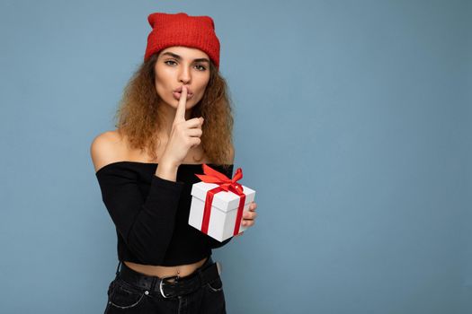 Attractive happy young dark blonde curly woman isolated over blue background wall wearing stylish black crop top and red hat holding gift box looking at camera and showing shh gesture.Copy space, mockup