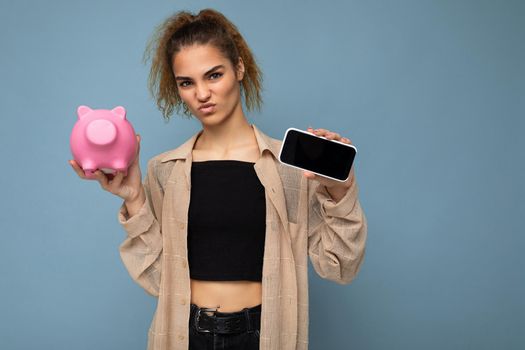 Photo of dissatisfied doubtful young beautiful attractive brunette curly woman with sincere emotions wearing stylish beige shirt isolated over blue background with copy space, holds pink pig penny bank for coins and shows smartphone with empty screen display. Payment concept.