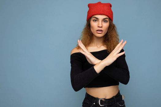 Shot of young amazed nice winsome brunet woman wavy-haired with sincere emotions wearing black crop top and red hat isolated on blue background with free space and showing stop gesture. Negative concept.